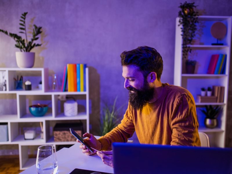 A happy millennial man is smiling at the phone while sitting in a purple and blue neon-lighted home office and purchasing online in a web shop. A cheerful man is spending money online.