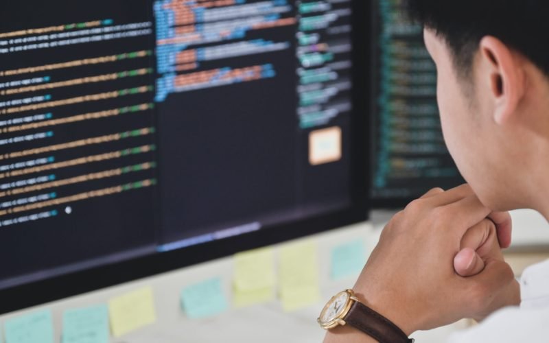 A male programmer sits and watches the code on a computer monitor to verify the program's execution.